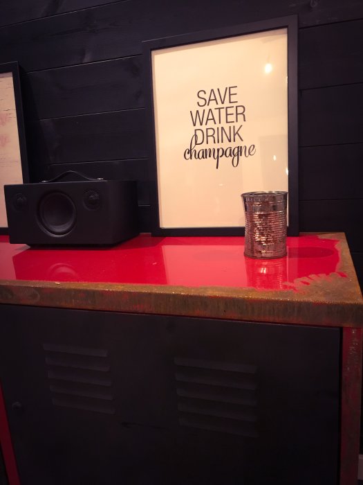Rostigt plåtskåp med röd topp, inramad affisch med texten "SAVE WATER DRINK champagne" och en högtalare.