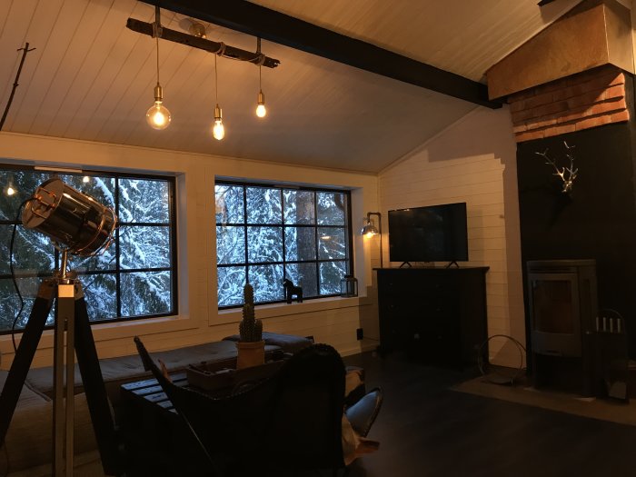 Rustikt renoverat vardagsrum med högt vitt tak, svarta detaljer och utsikt över snöbeklädda träd.