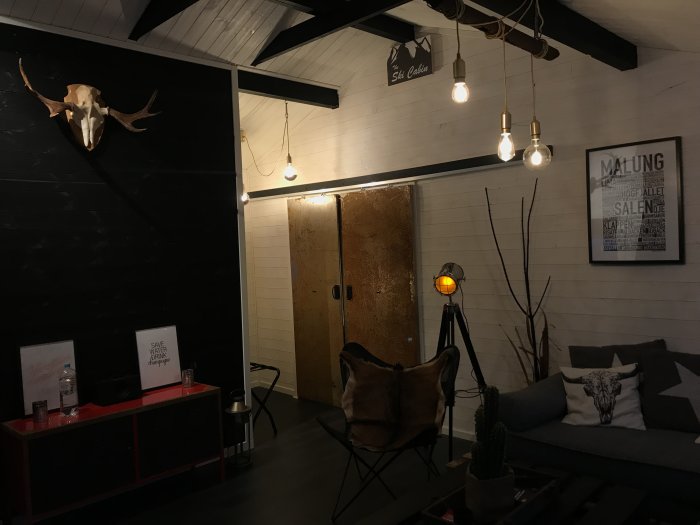 Rustikt inrett vardagsrum med svarta väggar, vit paneltak, industriell belysning, och vintage möbler.
