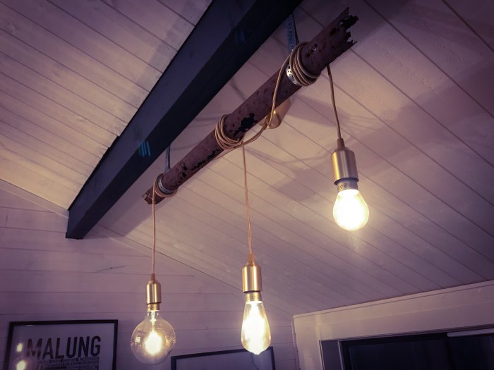 Hemmagjord lampa med rostigt rör och hängande glödlampor framför vit panelvägg och svart takbjälke.
