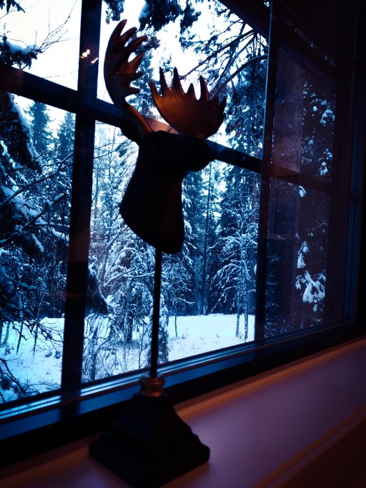 Dekorativ älgskulptur framför ett stort fönster som ger utsikt över ett snöigt skogslandskap i skymningen.