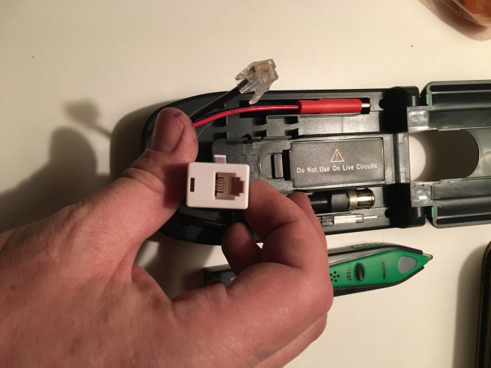 Hand håller en RJ45 adapter nära en öppen nätverkskabeltester med varningstext.