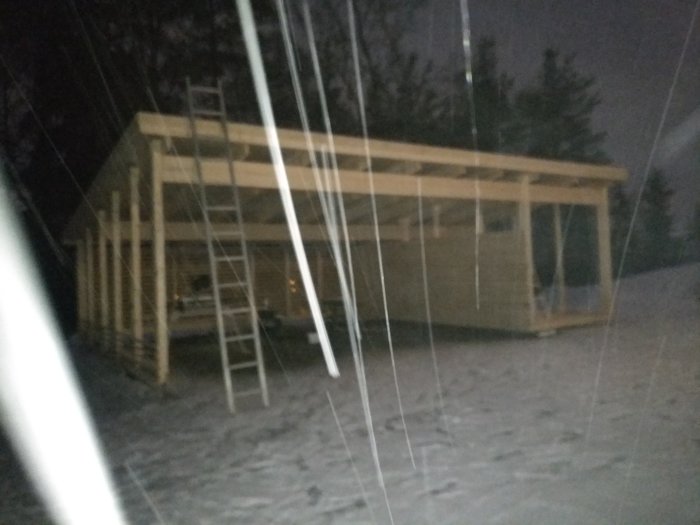 Byggkonstruktion av carport i kvällsbelysning under snöfall, med väggar delvis klara och omgivande snötäckt mark.