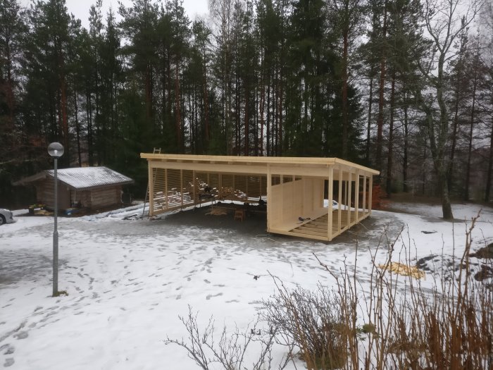 Halvfärdig träcarport med spjälor och påbörjat golv i snötäckt landskap.