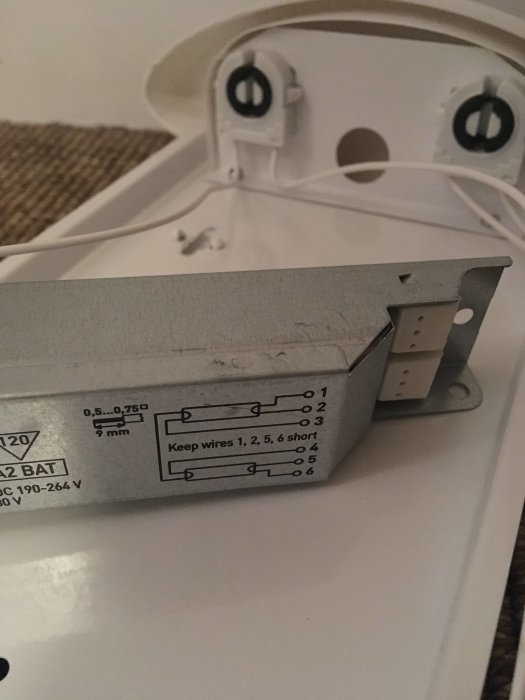 Närbild av ett elektroniskt drivdon med etikett och kopplingsschema, bredvid en vit lysrörsfäste.