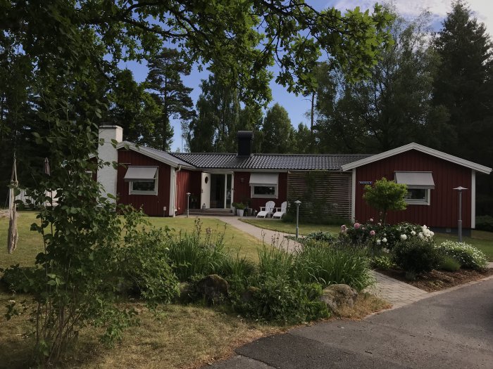 Ett rött hus och garage med vita knutar, omgivet av en trädgård med grönska och blommor.