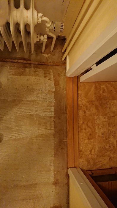 Hörnet av ett rum med en radiator, golvyta klar för parkettläggning och en fast list vid en dörrtröskel.