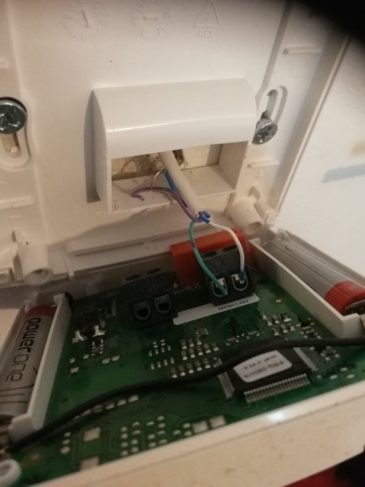 Öppet termostatfodral som visar en kretskort med anslutna turkosa och vita sladdar, mot en vägg.