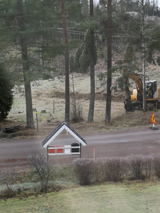 Utsikt över en byggarbetsplats med en minigrävare och en pittoresk postlåda i förgrunden, omgiven av skog och gräs.