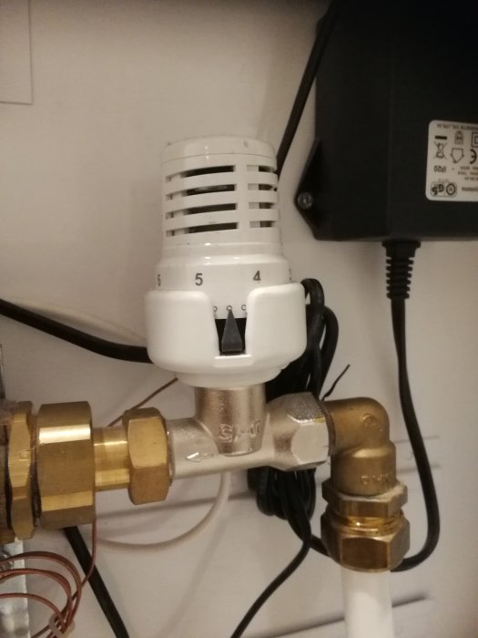 Termostatventil på radiator med inställningsskala och vita skyddslock ovanför ett rörkopplingssystem.