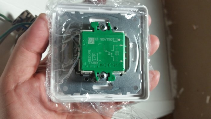 Hand håller en ny elektrisk strömställare med synliga kopplingsscheman, inuti plastförpackning.