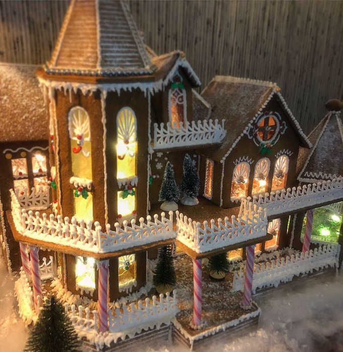Detaljerad pepparkakshus modell med ljus, verandor, staket och juldekorationer, belyst i mjukt ljus.