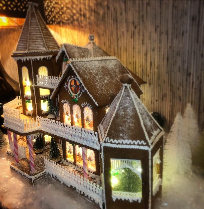 Modell av ett pepparkakshus med belysning och snödekoration bakom en vit staket.