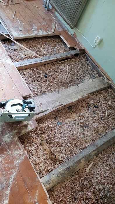 Delvis upprivet trägolv med synliga bjälkar och kutterspån, samt en cirkelsåg.