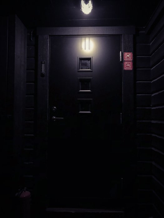Renoverad hall med ny svart ytterdörr, upplyst av en lampa ovanför och brandskyddsmärkningar vid sidan.
