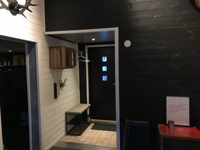 Renoverad hall med vit panel på väggarna, svart ytterdörr, toalettdörr med frostat glas och ett laminattak som liknar betong.