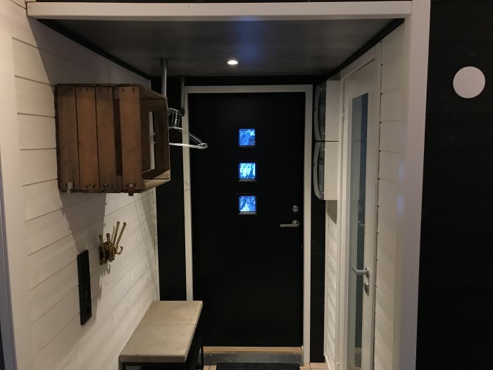 Renoverad hall med vit panelvägg, svart ytterdörr med fönster och laminattak som efterliknar betong.