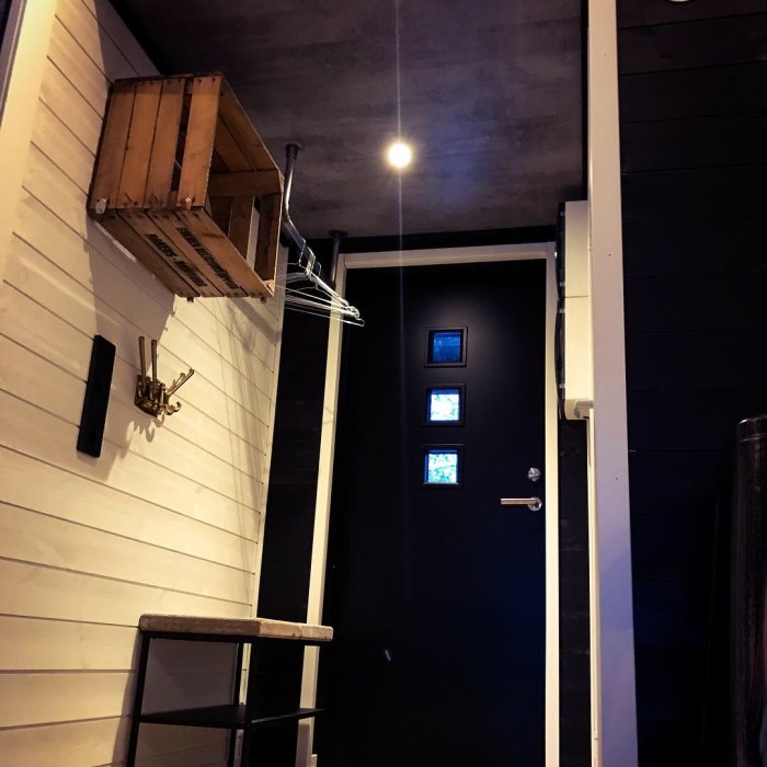 Renoverad hall med svart ytterdörr med fönster, vit panelvägg och mörkt laminattak efterliknande betong.