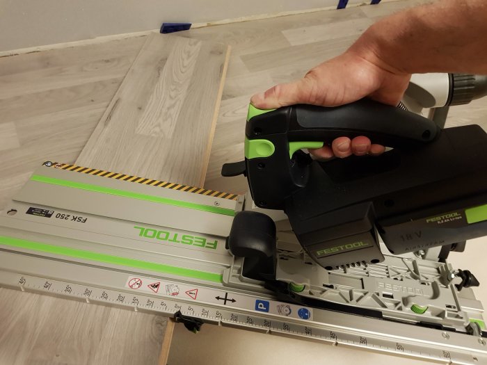 Hand som använder en Festool HKC 55 sladdlös cirkelsåg på en skena för att kapa laminatgolv.