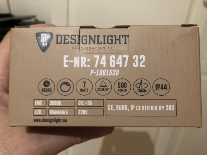 Hand håller en kartong från DESIGNLIGHT med produktspecifikationer för en dimbar LED-lampa, 590 lumen, 3000K.