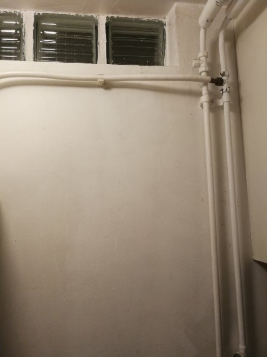 En vitmålad tvättstugerör med gamla synliga rör längs väggen och i taket nära ett par frostat glasfönster.