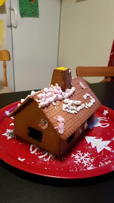 Pepparkakshus dekorerat av barn med godis och strössel på ett rött fat, snöflingedekorationer, visst renoveringsbehov.