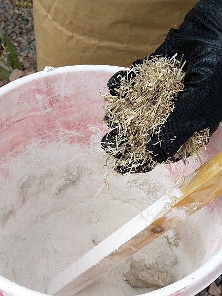 Hand i svart handske blandar klippt linhalm i en hink med kalkbruk och murarslev.