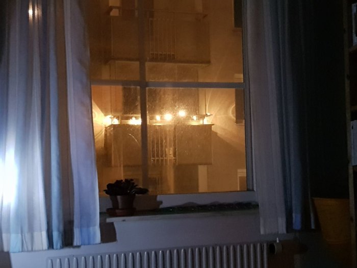 Mörk interiörbild med fönster som visar reflektion av ett tänd ljus, gardiner och en blomma på fönsterbräda.