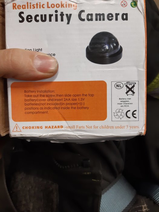 Hand håller en kartong för en "Realistic Looking Security Camera" med felstavad varningstext "CHOKING HAZARD - small Farts".