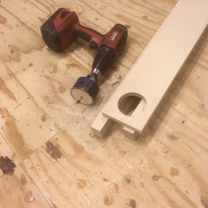 Träbit med utborrat hål bredvid en borr och hålsåg på ett sågspånat bord.