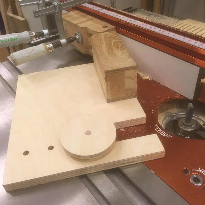 Verkstadsscenario med en delvis konstruerad jig, en klemm och träbitar på ett sågspånbelagt fräsbord.