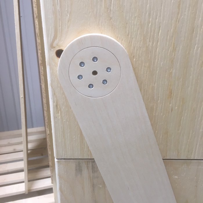 Trädetalj med flera skruvar och borrade hål, troligen en del av en länkarmskonstruktion.