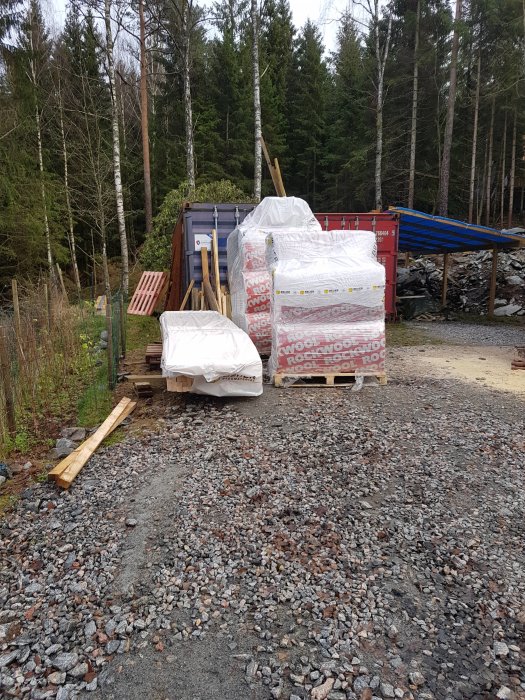 Isolermaterial på pallar vid byggarbetsplats med skog i bakgrunden.
