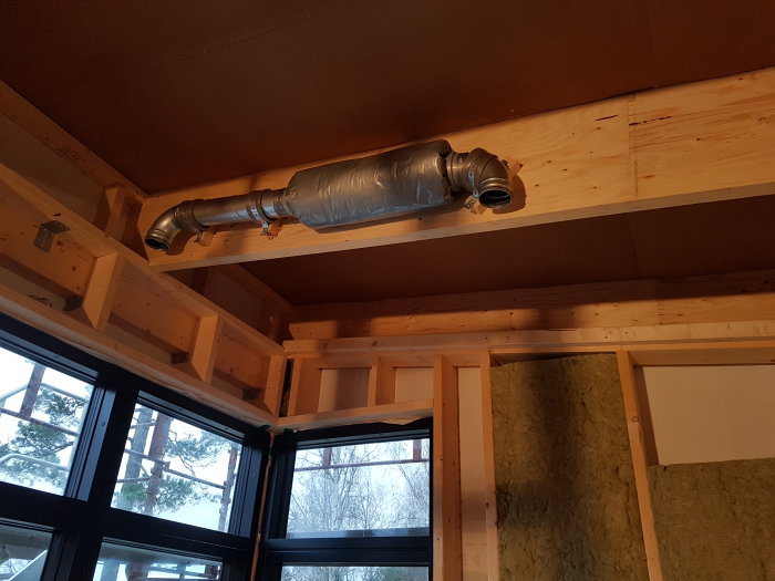 Ventilationsrör monterat mellan träbalkar i tak, omgiven av isolering och plastfolie.