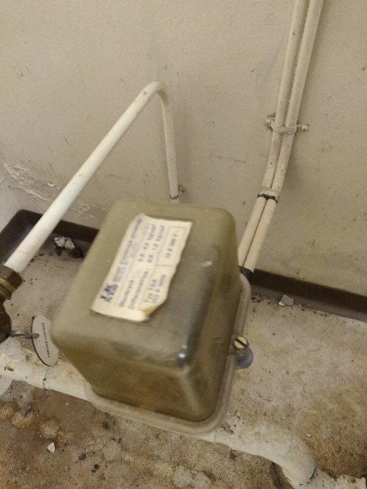 En brun vattenmätare kopplad till vit rörledning med etikett, installerad vid en betongvägg.