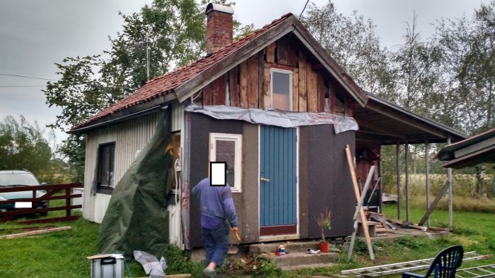 Hus med obeklädd fasad och synligt regelverk under renovering, tak med röda tegelpannor, person framför huset.