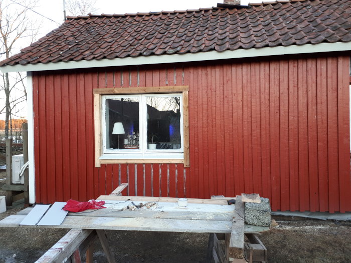 Rött trähus under renovering med öppet fönster och byggmaterial framför.