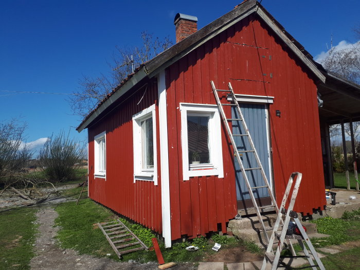 Rödmålat hus i början av renovering med stege lutad mot väggen under en klarblå himmel.