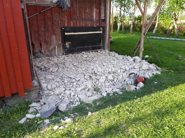 Grushög framför gammal röd ladugård, skottkärra och sladdborrmaskin i förgrunden.