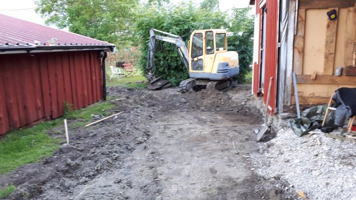 Grävmaskin vid arbete i trädgården nära rött uthus, med grushög och grävd mark.