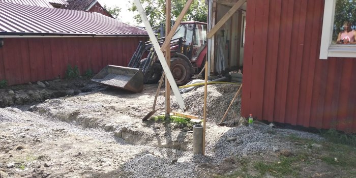 Grundarbete för byggprojekt med grävmaskin och grus nära röd stuga.