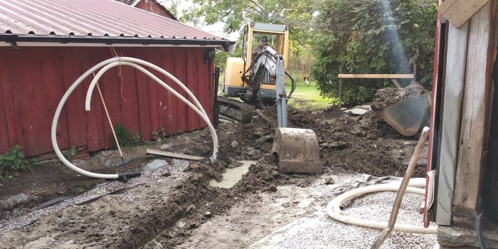 Grävmaskin arbetar med att gräva ur marken bredvid röd träbyggnad, jord och slangar syns.