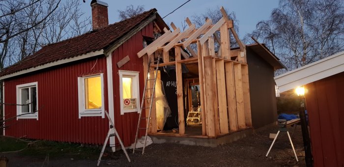 Renovering av röd stuga med pågående tillbyggnad i träkonstruktion i skymningen.