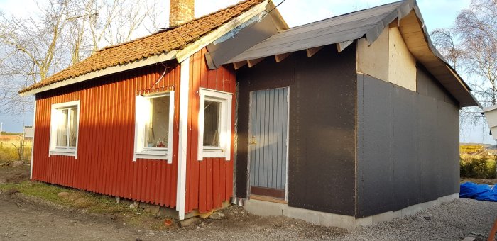 Rödfärgad stuga under renovering, saknar panel på tillbyggnad och beredd för fler fönster och altandörr.