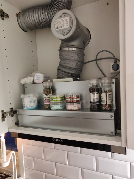 Flexibel ventilationskanal kopplad till mätare över en köksfläkt med kryddburkar framför vit kakelvägg.