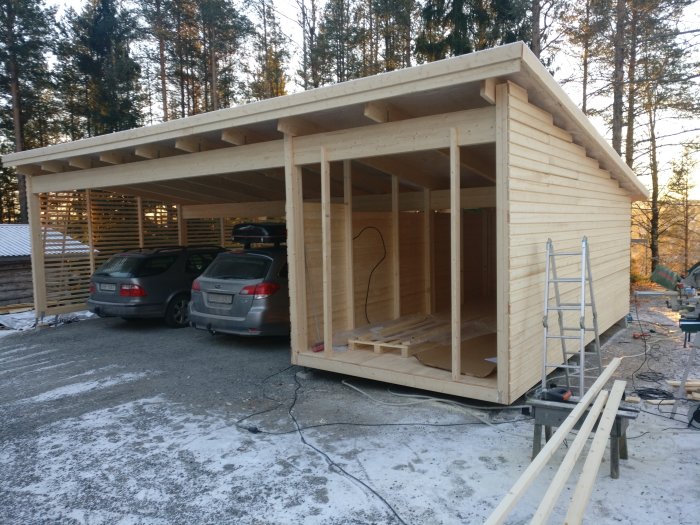 Nästan färdigbyggd trädgårdsstuga med träpanel och inbyggd carport, verktyg och bilar syns.