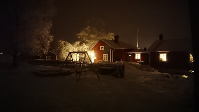 Vinterkväll med snötäckt trädgård och upplyst rött hus vid bastubad.