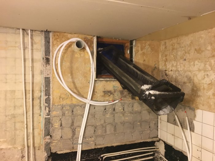 En svart takränna som står på snedden i ett ouppvärmningsbart utrymme med synliga rör och murat väggmaterial.