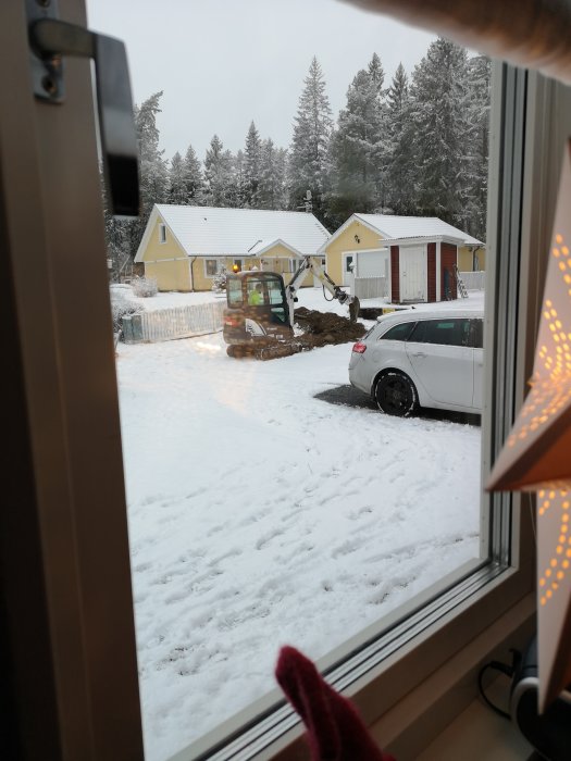 Minigrävare som gräver nära en snötäckt uppfart, sett från ett fönster, med ett vitt hus och skog i bakgrunden.