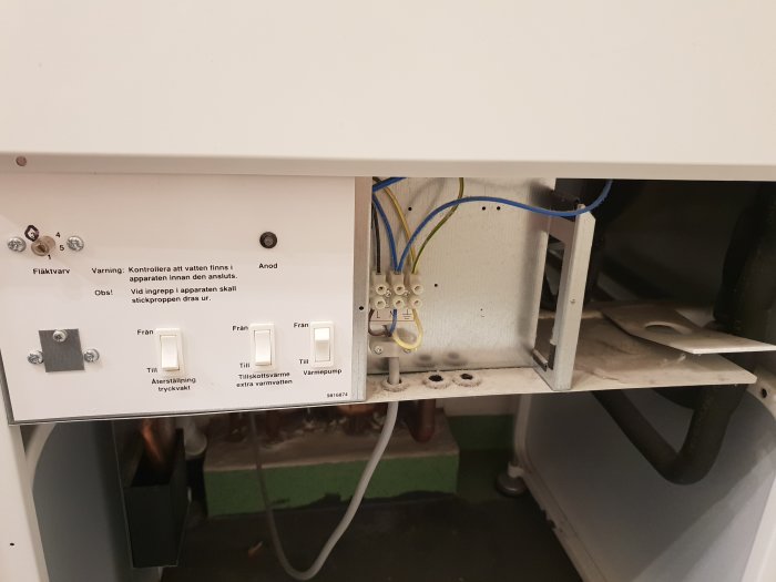 Öppen insida av ett värmesystem med ledningar, etiketter och bortskruvad plåt.